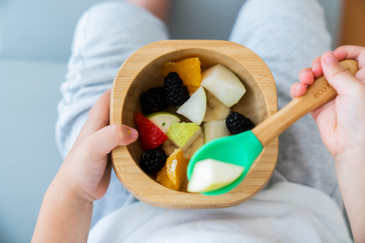 Criança a segurar na tigela de bambu com frutas frescas variadas e colher de silicone verde