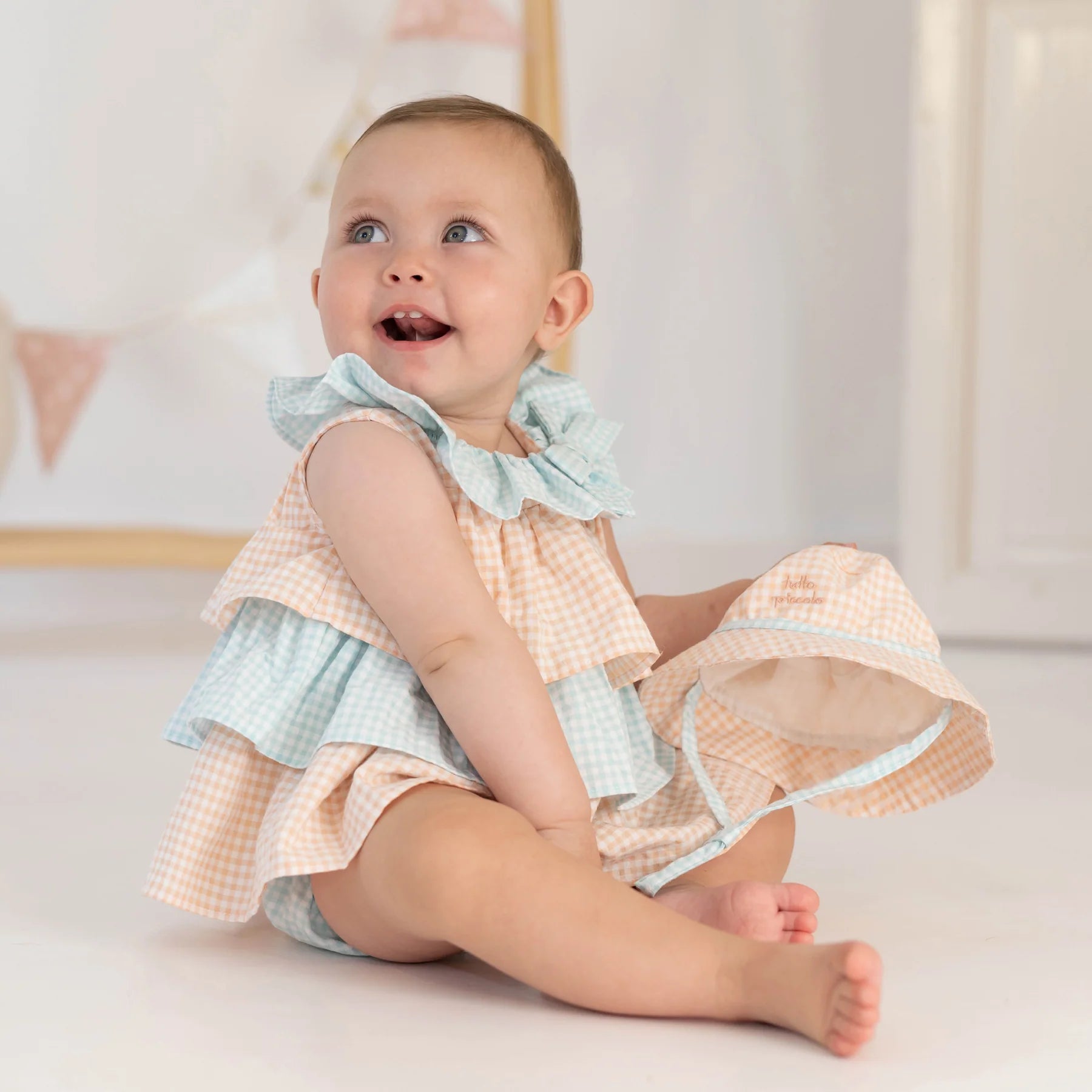 Bebé sorridente a usar o vestido xadrez pêssego/menta da Tutto Piccolo com tapa-fralda, segurando um chapéu a condizer.