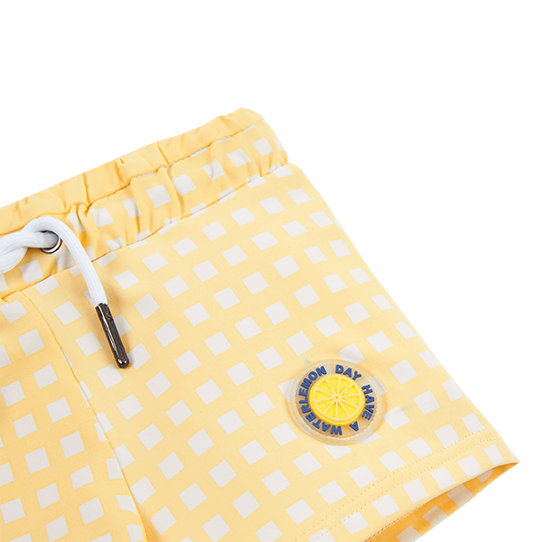 Detalhe do padrão xadrez amarelo e branco e do emblema redondo com limão no calção de praia Waterlemon.