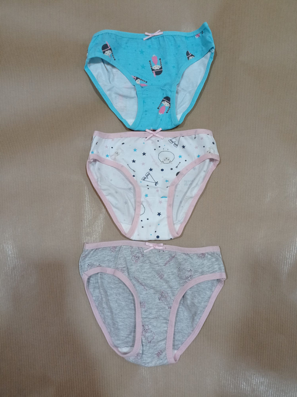 Três cuecas para criança, em tons azul, branco e cinzento, sobrepostas numa superfície castanha. As cuecas mostram detalhes das estampas.