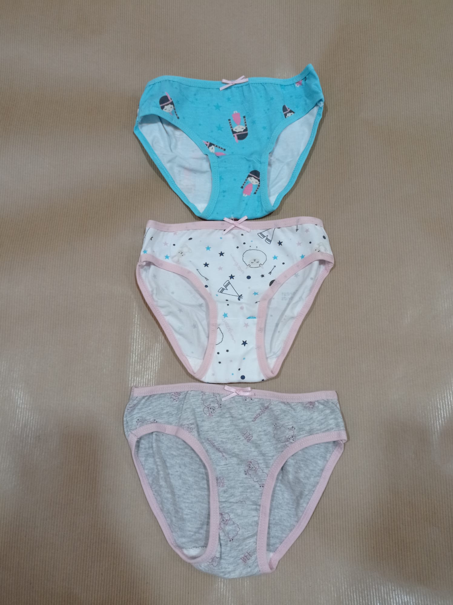 Três cuecas para criança, em tons azul, branco e cinzento, sobrepostas numa superfície castanha.
