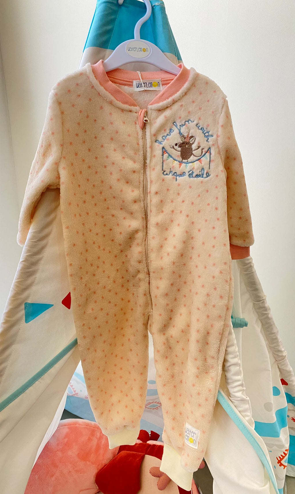 Pijama macacão Waterlemon rosa claro com padrão de bolinhas e um rato bordado no peito, pendurado num cabide contra um fundo de tenda infantil.