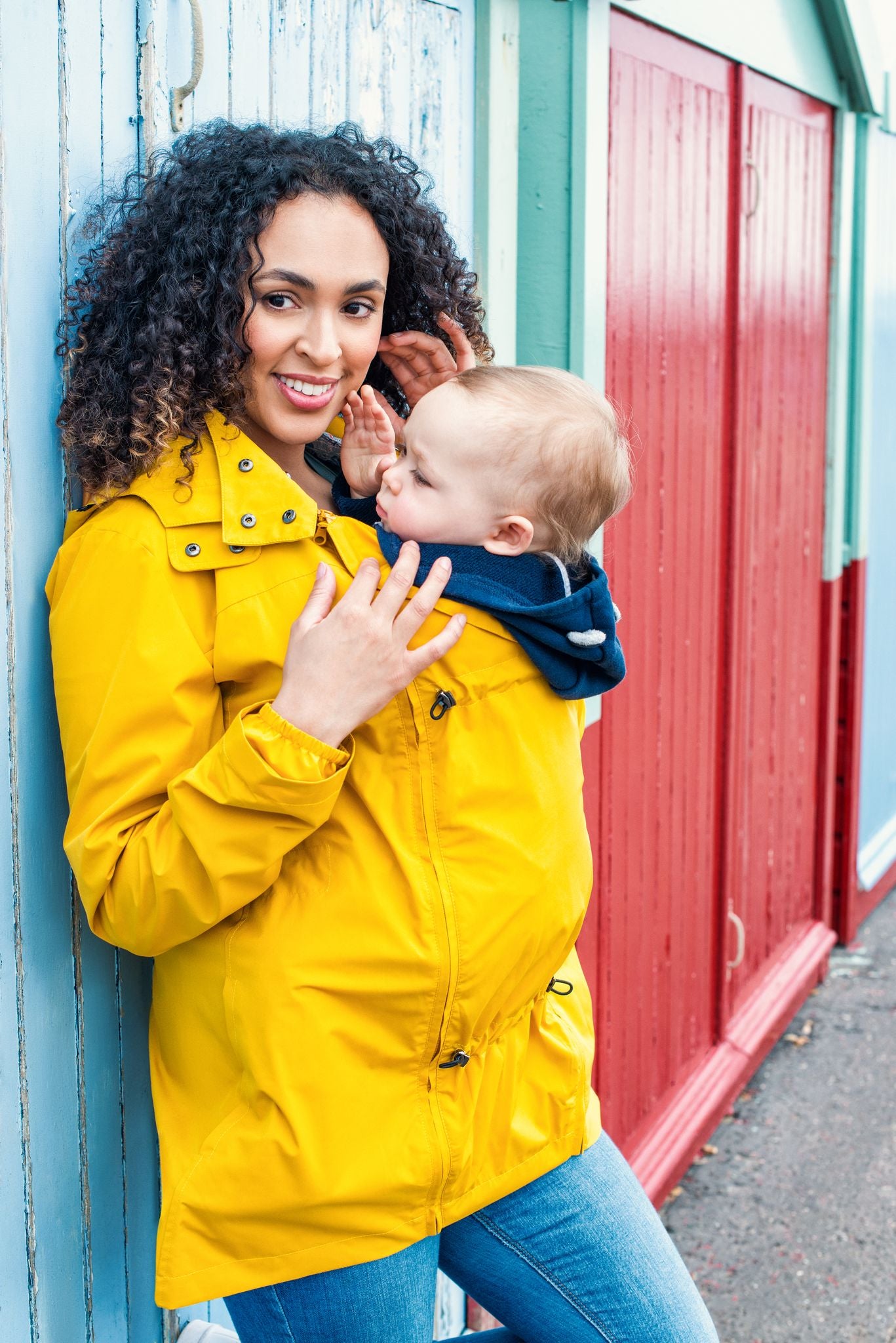 Mãe sorridente com cabelo encaracolado usa o casaco amarelo Wombat Numbat GO para transporte de bebé à frente.
