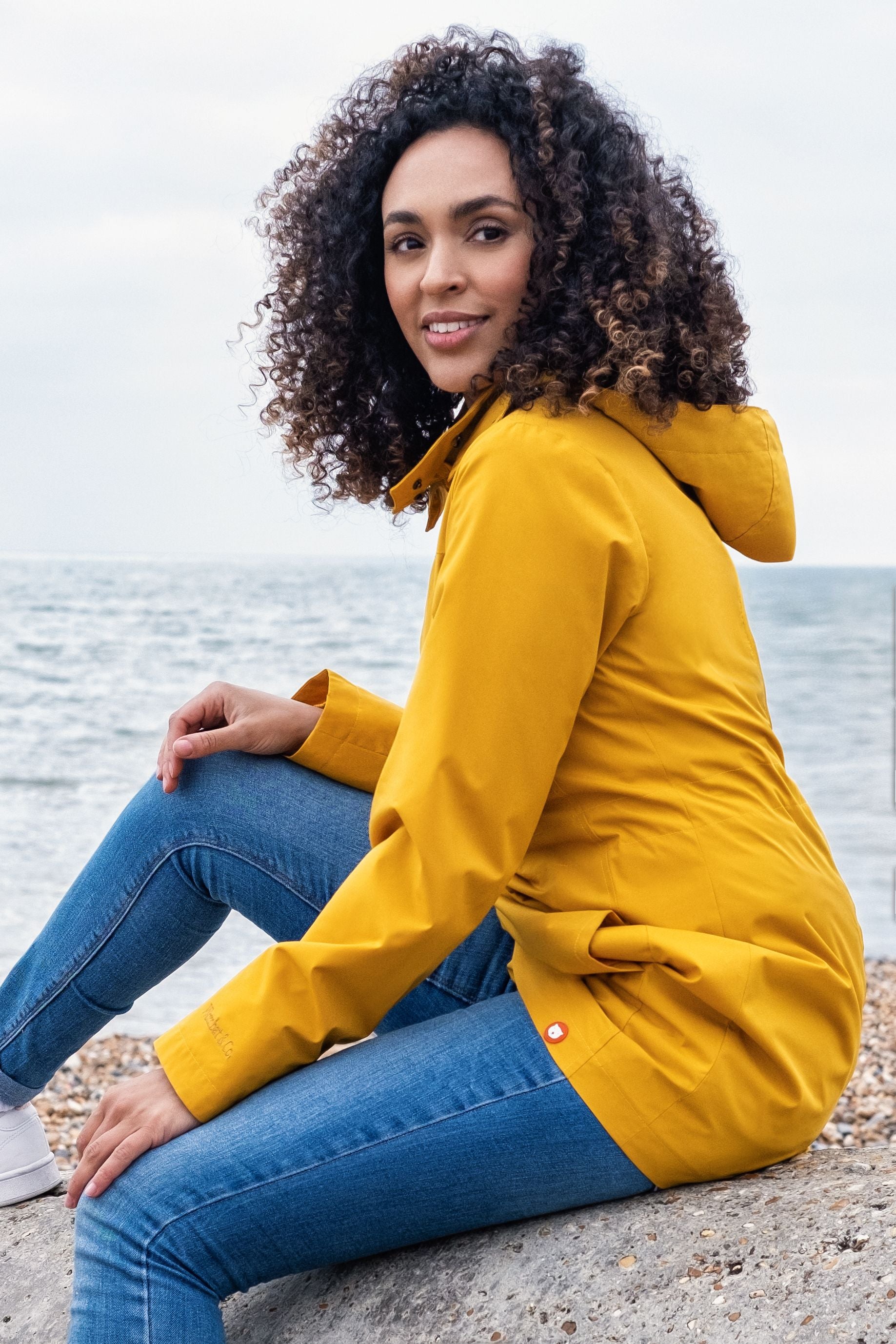 Mulher com casaco Wombat Numbat GO amarelo sentada numa praia, com o mar ao fundo, em pose casual.