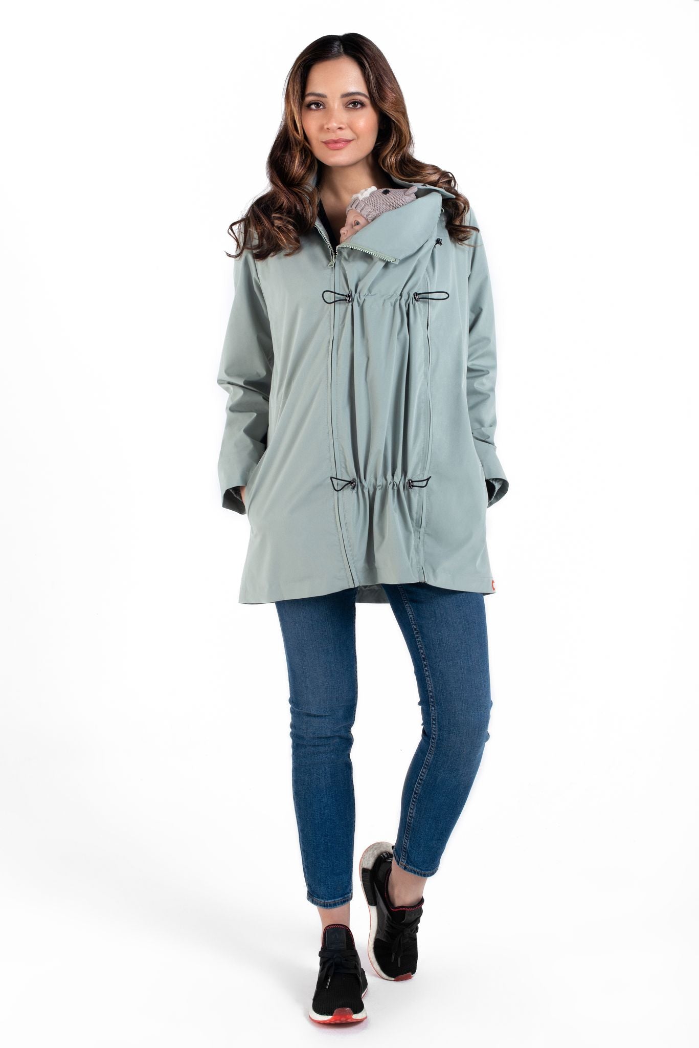 Mulher sorridente a usar o casaco Wombat Numbat GO 4 em 1, ajustado para transporte de bebé à frente, cor verde-água.