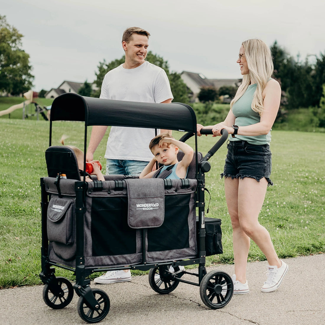 Família feliz usa Wonderfold Wagon Serie W para 2 crianças em passeio no parque. Carrinho vagão confortável e seguro.