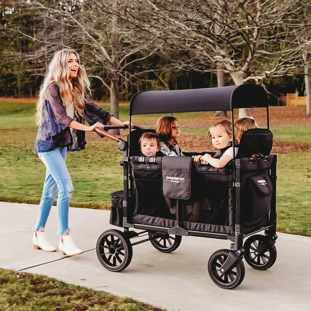 Mãe empurrando Wonderfold Wagon Serie W preto com 4 crianças no parque. Carrinho vagão versátil para família grande.