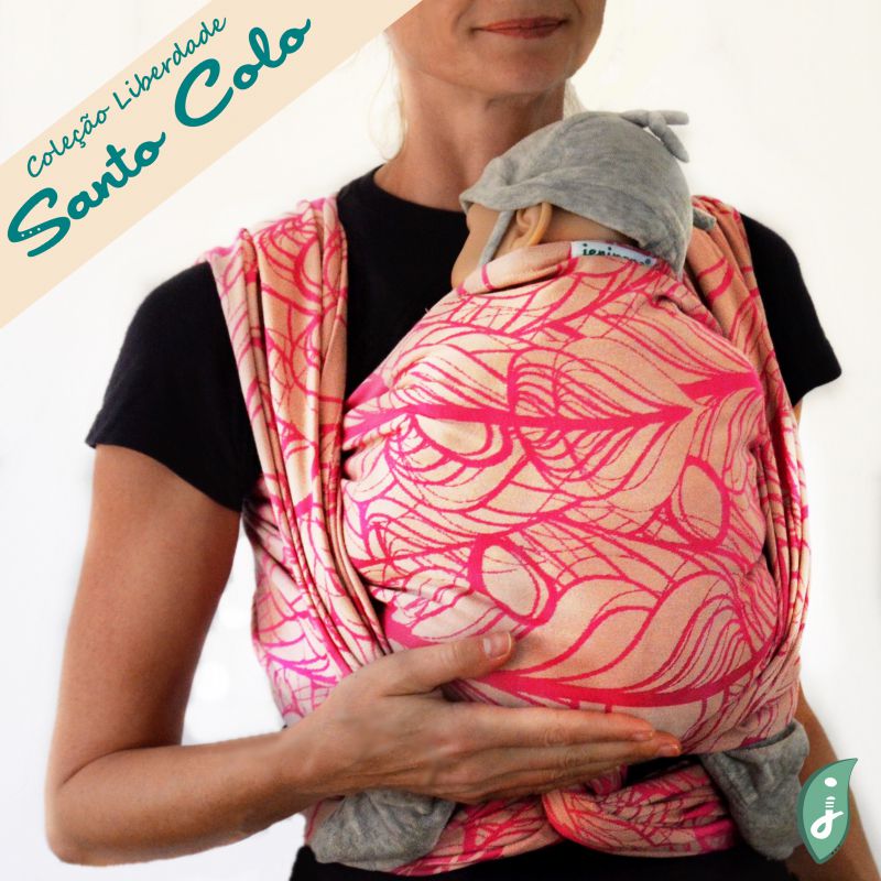 Mulher usando um pano tecido da coleção Liberdade de Santo Colo, na cor rosa com padrões orgânicos, carregando um bebé. O pano é envolto de forma segura e confortável, mostrando a versatilidade e elegância do design.